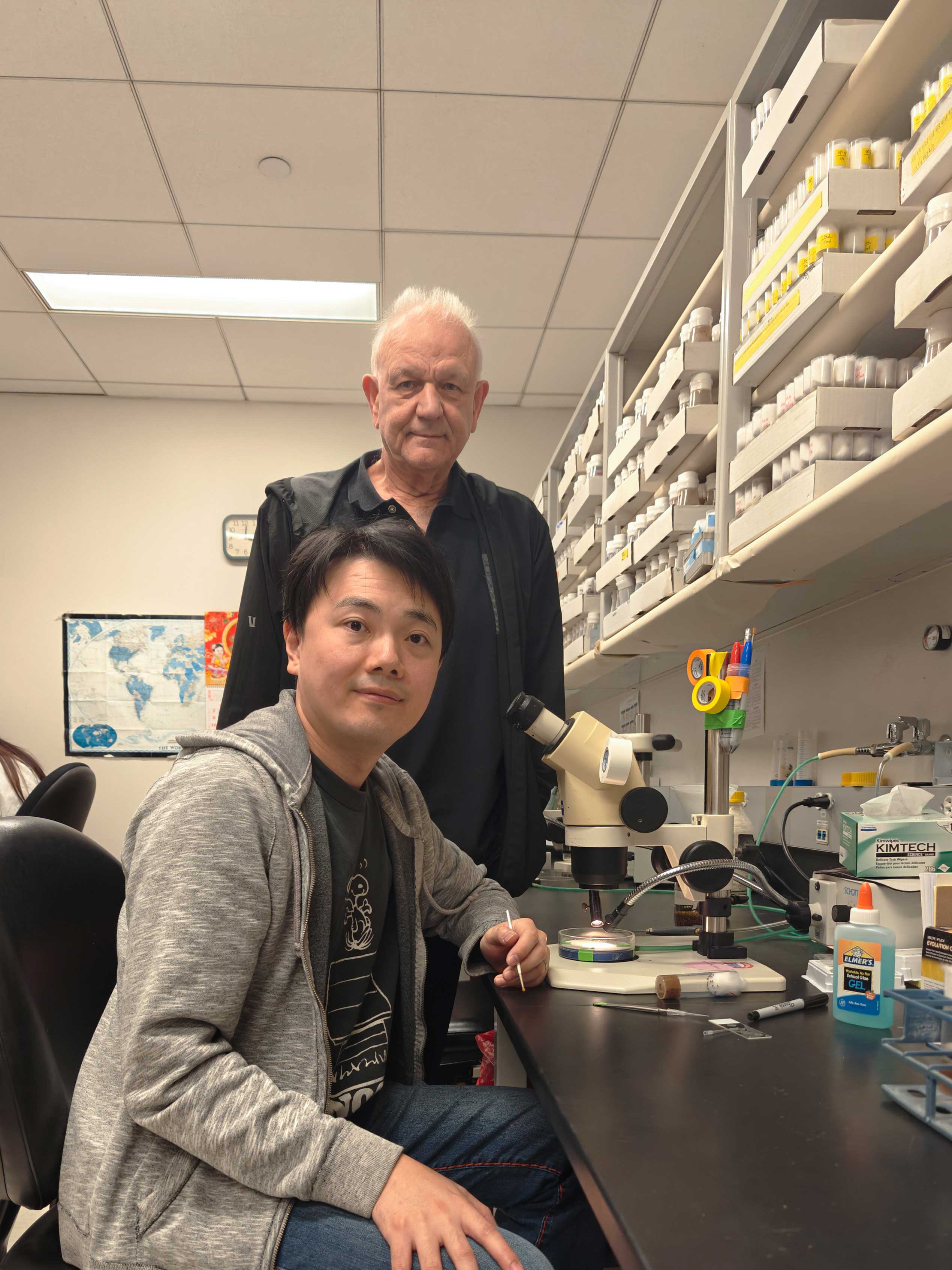 Xueyang Pan and Hugo Bellen in front of a dissection scope in a laboratory.